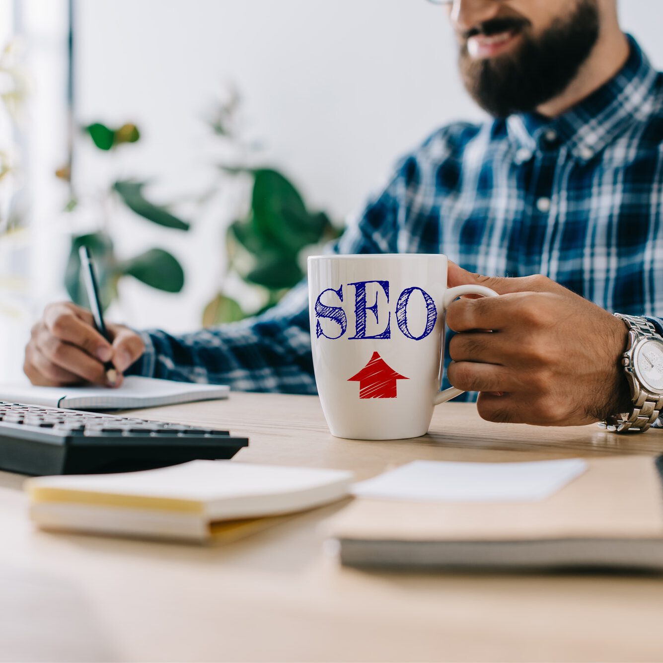 cropped shot of successful smiling seo developer with cup of coffee and computer writing notes at